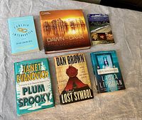 Photo showing a collection of 7 assorted books including 2 National Geographic hard cover photography and nature books and 5 fiction novels in paperback form.