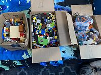 Three large cardboard boxes filled with assorted Lego pieces and parts, including loose bricks and bagged items
