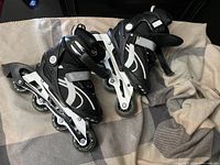 Pair of black and white youth size 5 roller blades with four wheels inline, adjustable straps, placed on a grey and beige checkered fabric.