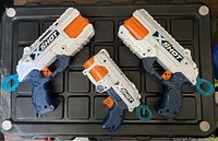 Three X Shot Nerf toy blasters displayed on black padded surface, two large and one small, all white and gray with orange and blue accents, no darts included.