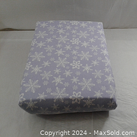 Folded box spring fitted sheet showing light grayish-purple fabric decorated with white snowflakes in various sizes.