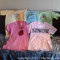 Five girls T-shirts laid out flat showing front designs: yellow with flower, light blue with dolphin, light green with cartoon tiger, bright pink with ladybug and 'REBEL', light pink with black bird silhouettes.