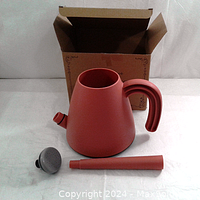 Terracotta-colored plastic watering can jug with curved handle, detached long spout and removable spray head placed in front of its cardboard box.