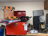 Full view showing red Skil metal box open with belt sander inside, sandpaper belts, and adjacent tools in metal cases and on table.