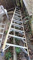 Full length view of a long fiberglass ladder with aluminum rungs, showing black rubber feet at one end and dirt on the ground beneath it.