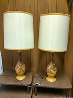 Pair of ceramic base lamps with mottled brown and yellow glazed bases on wooden tables under off-white drum shades with yellow trim.