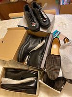 Photo showing multiple shoes in boxes including black lace-up boots, black polished dress shoes, and brown plaid slippers.