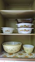 Photo of vintage Pyrex and Fire King bowls and casserole dishes arranged on shelves, showing rooster pattern on Fire King dishes and Wintergreen floral pattern on Pyrex bowls