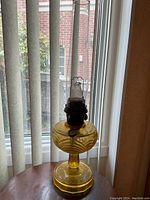 Full view of the antique amber glass Aladdin oil lamp with chimney standing on wooden surface in front of window with vertical blinds.