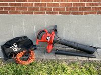 Photo showing the full Black and Decker Leaf Hog blower/vacuum with orange extension cord, black storage bag, and two black plastic tubes laid out on ground against a brick wall.
