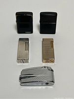 All five lighters displayed together: two black Zippo, two patterned slim lighters (one gold, one silver), and one chrome silver lighter with curved design