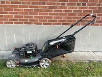 Side view of the Remington gas powered lawn mower with grass collection bag attached, positioned on grass near a brick wall.
