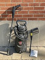 Overall view of JobMate 1500 PSI electric pressure washer unit, two spray gun attachments, and instruction manual on stone surface against a brick wall.