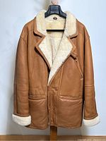 Front full view of men's tan shearling coat hanging on a hanger showing tan leather and cream shearling lapels and cuffs.
