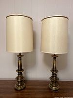 Pair of vintage brass tone metal table lamps with cream fabric drum shades sitting on wooden furniture against white wall. Both lamps shown fully assembled.