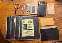 Photo shows all stamp collecting supplies arranged on a wooden surface including protective sleeves, mount sheets, tools like magnifying glass and tongs, mint block booklet and envelopes.