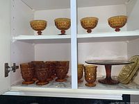 View of amber glass dessert glasses on upper shelf, large and small drinking glasses and cake platter on lower shelf.