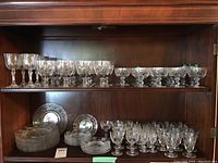 Wide view of cabinet shelves showing assortment of crystal stemware glasses on the top two shelves and matching plates on the bottom shelf. Variety of shapes and sizes, some with intricate cut patterns. Green stickers visible on some items indicating quality control or inventory tagging.