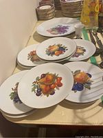 Twelve porcelain dessert plates and two serving plates with fruit designs stacked, showing various fruit images on white tableware with gold rims. One porcelain egg cup is seen near the serving plates.