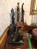 Six figurines displayed on a wooden surface against a white textured wall. Includes a bronze-colored kneeling figure, wooden elephant, painted female figure, wooden abstract human, and two elongated dark metal sculptures.