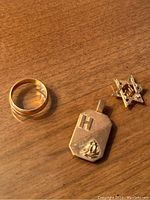 Photo showing a gold band ring, a 10k gold rectangular medallion pendant marked with an H and figure, and a 14k Star of David pendant with Hebrew script.