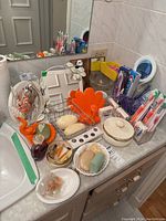 Overview of health and beauty products laid out on bathroom counter including toothbrushes, razors, soaps, perfume samples, mirrors, and soap dishes.