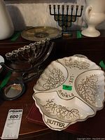 Two menorahs, divided ceramic dish with gold outlined leaf and grape decorations labeled for Lox, Bagels, Butter, Cheese, and a silver-tone round coaster with Hebrew inscription visible on the edge.
