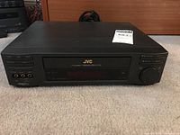 Front view of black JVC HR-VP1605U VHS player on carpet, showing control buttons, VHS slot, and input jacks