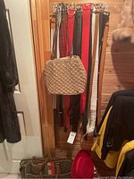 View of multiple ladies belts hanging on rack featuring various colors and styles including woven, leather, and synthetic materials. Beige woven purse also visible attached to rack, along with multicolor handbag and red hat on floor below.