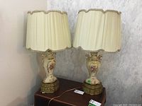 Pair of ceramic table lamps with floral painted urn-shaped bodies, gold-tone ornate metal bases, and pleated cream fabric lampshades with decorative trims sitting on a wooden table against a textured wall.