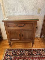 Front view of wood nightstand showing one drawer with brass handle and lower cabinet with two doors and brass knobs, topped with a glass pane.