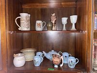 Two shelves showing a variety of pottery items including mugs, vases, teapot, honey pot, and goblets in a wooden cabinet.