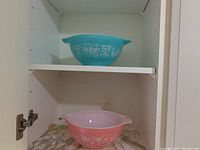 Two Pyrex bowls, one turquoise blue with white floral and figure patterns, and one pink with white floral patterns, placed on shelves of a white cabinet.