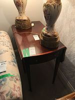 Side view of the dark wood side table showing the single drawer and one dropped leaf folded down, placed against a wall with lamps on top.