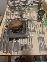 Full view of silver plated cutlery set arranged on table with silver serving tray, bread basket, lidded box, and salt and pepper shakers.