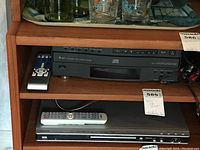 Sony 5-disc CD player shown on wooden shelf with remote control on left side of the unit. Several drinking glasses and bottles are on the shelf above.