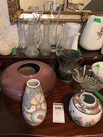 Photo shows assortment of glass, metal, and porcelain vases and pots on a wooden surface in front of a mirror.
