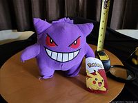 Front view of the Gengar plush showing its facial features, size approximate by measuring tape, and attached Pokémon tag.