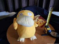 Front view of the Psyduck plush toy, showing face with white eyes and large beige beak, alongside measuring tape showing approximately 10 inches.