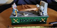 Side view showing two hockey players and a goal on a cast-iron coin bank with HOCEKY and crossed sticks on front.