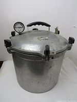 Full view of pressure cooker showing aluminum body, multiple black clamp handles securing the lid, pressure gauge, and black handle on lid.