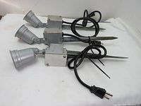 Photo showing three metal waterproof outdoor spot lights with attached power cords and two-prong plugs, arranged horizontally with stakes visible.