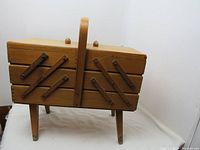 Front corner view of vintage wooden accordion sewing box on legs with handle on top. Shows accordion wood supports and legs