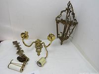 Photo displaying both items including the brass wall candle holder lying flat and the brass hanging lamp angled upright showing the decorative details and bulb sockets.