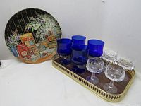 Set of vintage Italian mid-century serving trays and glasses including cobalt blue and white liquor glasses on an ornate brass tray with a wooden base, and a round decorative metal tray featuring vintage pantry graphics.