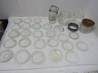 Overhead photo showing 25 glass canning jar lids arranged in rows alongside four vintage glass jars including one with cork lid and metal screw band.