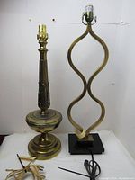Two vintage metal table lamps shown side by side on a white surface with plain white background. The lamp on the left has a traditional solid brass base with vertical grooves and decorative details, standing 24 inches tall. The lamp on the right has a modern artistic open frame curved metal design resting on a black square base, standing 27.5 inches tall. Both lamps have cord plugs visible and no bulbs installed.