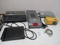 Vintage office equipment including Sanyo dictating machine with foot pedal, video cassette rewinder, yellow letter opener/pencil sharpener, Energizer battery charger, and cables on table.