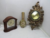 Photo showing a vintage ornate wall clock with gold and black dial, a wood Bulova quartz table clock, and a brass Taylor barometer with mounted thermometer; items displayed against white background.