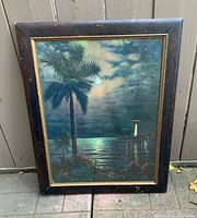 Front view of vintage framed lithograph showing tropical island scene with lighthouse and palm trees at night, with moonlight reflecting on water.